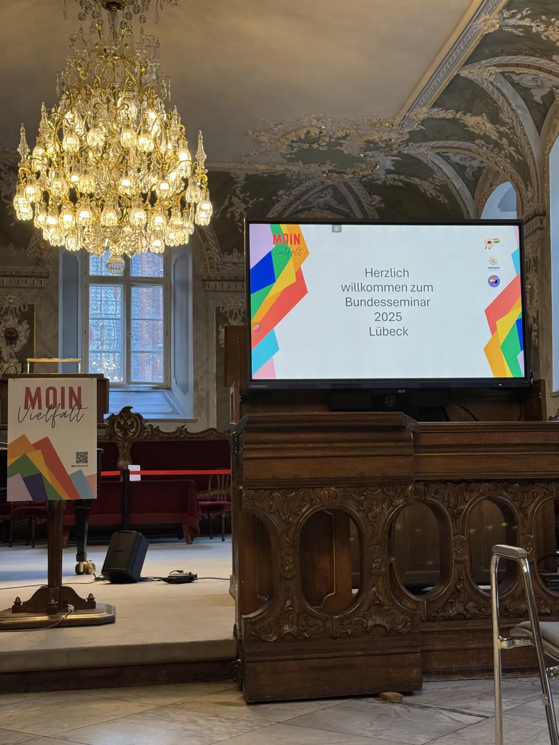 Blick auf die Bühne im Festsaal: ein Aufsteller mit dem Motto MOIN Vielfalt neben der Leinwand mit der Begrüßung zum Bundesseminar 2025.