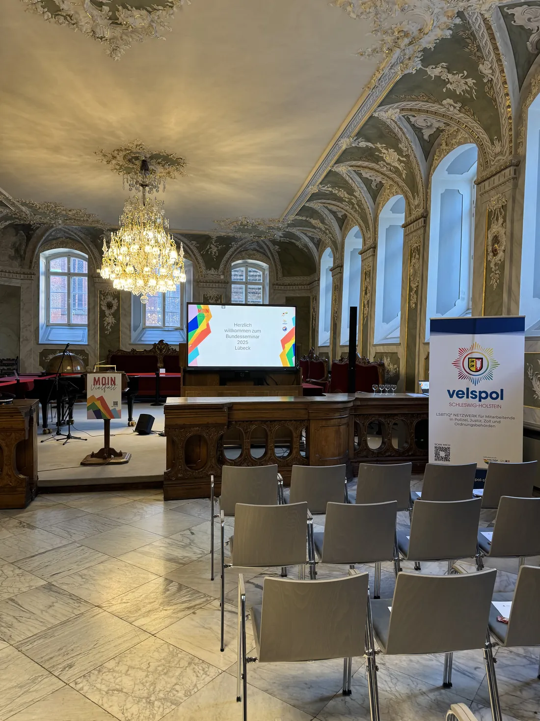 Der Festsaal des Lübecker Rathauses mit Stuckdecke und Kronleuchter, Stuhlreihen vor der Bühne und einem velspol-Rollup.