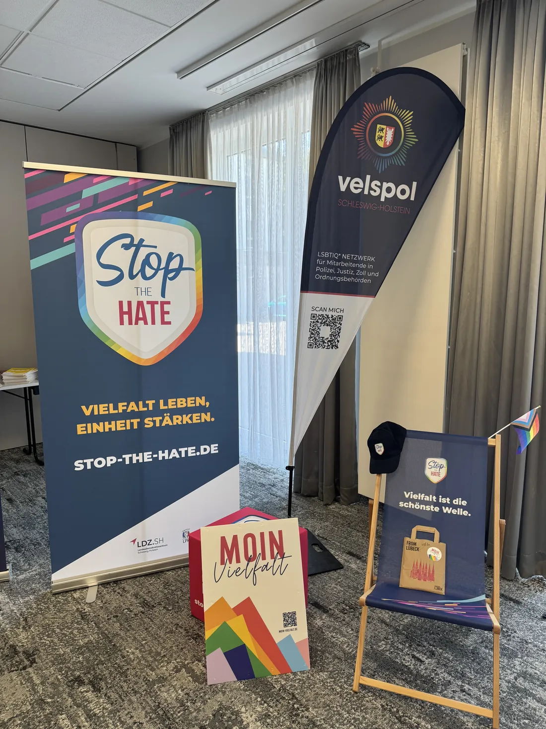 Aufsteller im Tagungsraum: STOP the HATE, eine velspol-Beachflag und das Motto MOIN Vielfalt neben einem Liegestuhl.